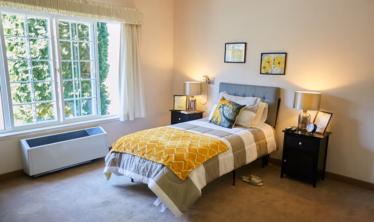 A cozy bedroom with a single bed featuring a gray and white comforter and a yellow throw blanket. The bed has decorative pillows and is flanked by two black nightstands, each with a lamp and framed pictures. A large window with white curtains lets in natural light, and a pair of slippers is placed on the carpeted floor beside the bed.