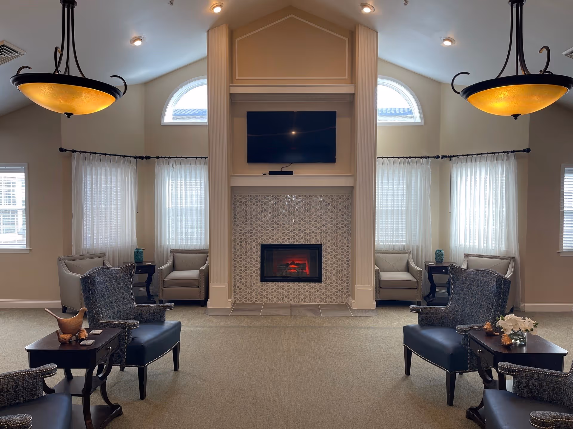 A cozy living room area with a central fireplace featuring a patterned tile surround and a mounted flat-screen TV above it. The room has high ceilings with two arched windows on either side of the fireplace and sheer white curtains on the windows. There are multiple armchairs arranged around small dark wood tables, with decorative items such as a wooden bird sculpture and a small vase with white flowers. Two pendant lights with warm yellow shades hang from the ceiling.