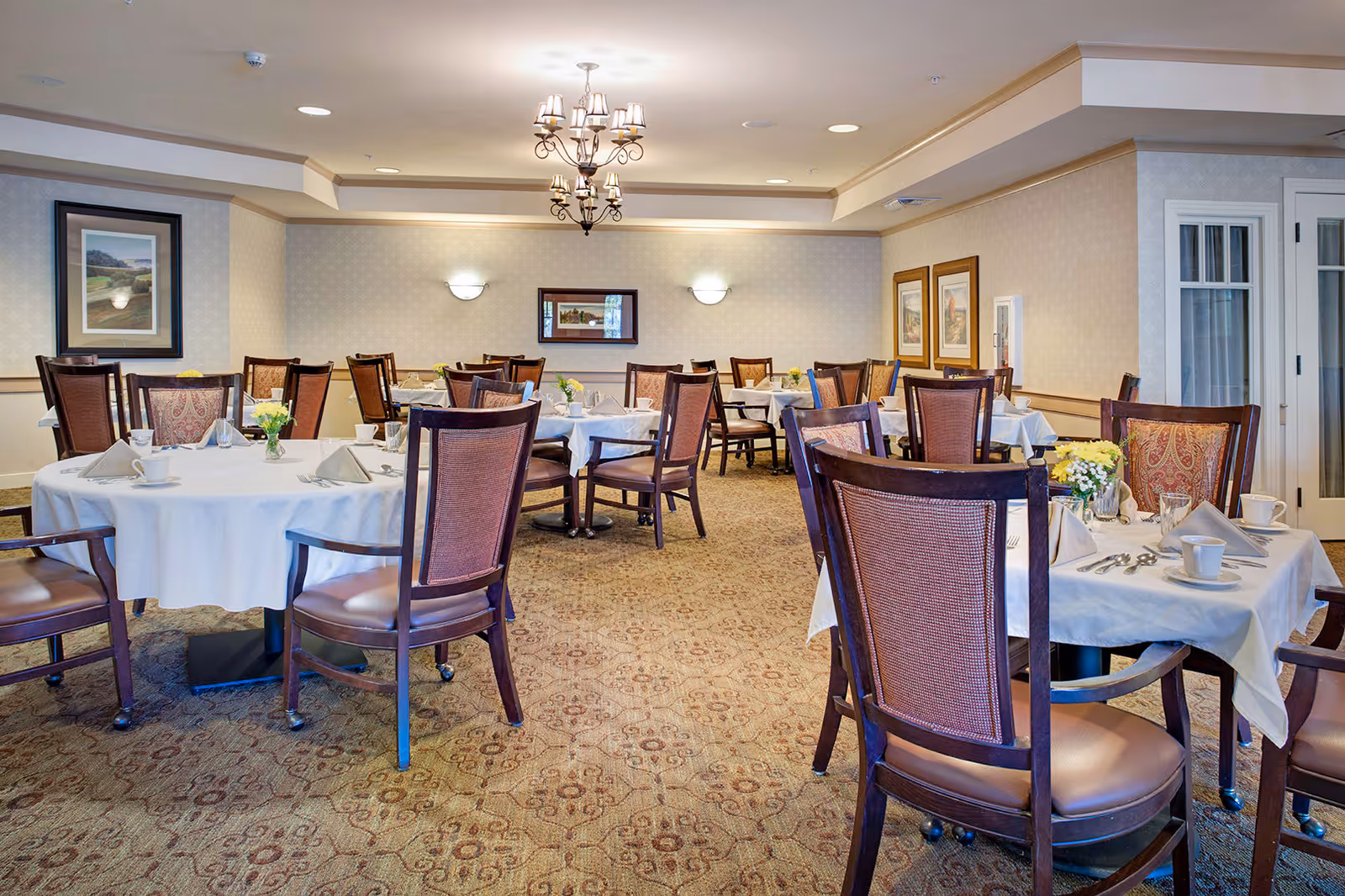 A well-lit dining room with multiple round tables covered with white tablecloths, each set with cups, napkins, and silverware. The room features wooden chairs with cushioned seats and backs, a patterned carpet, framed artwork on the walls, and a chandelier hanging from the ceiling.