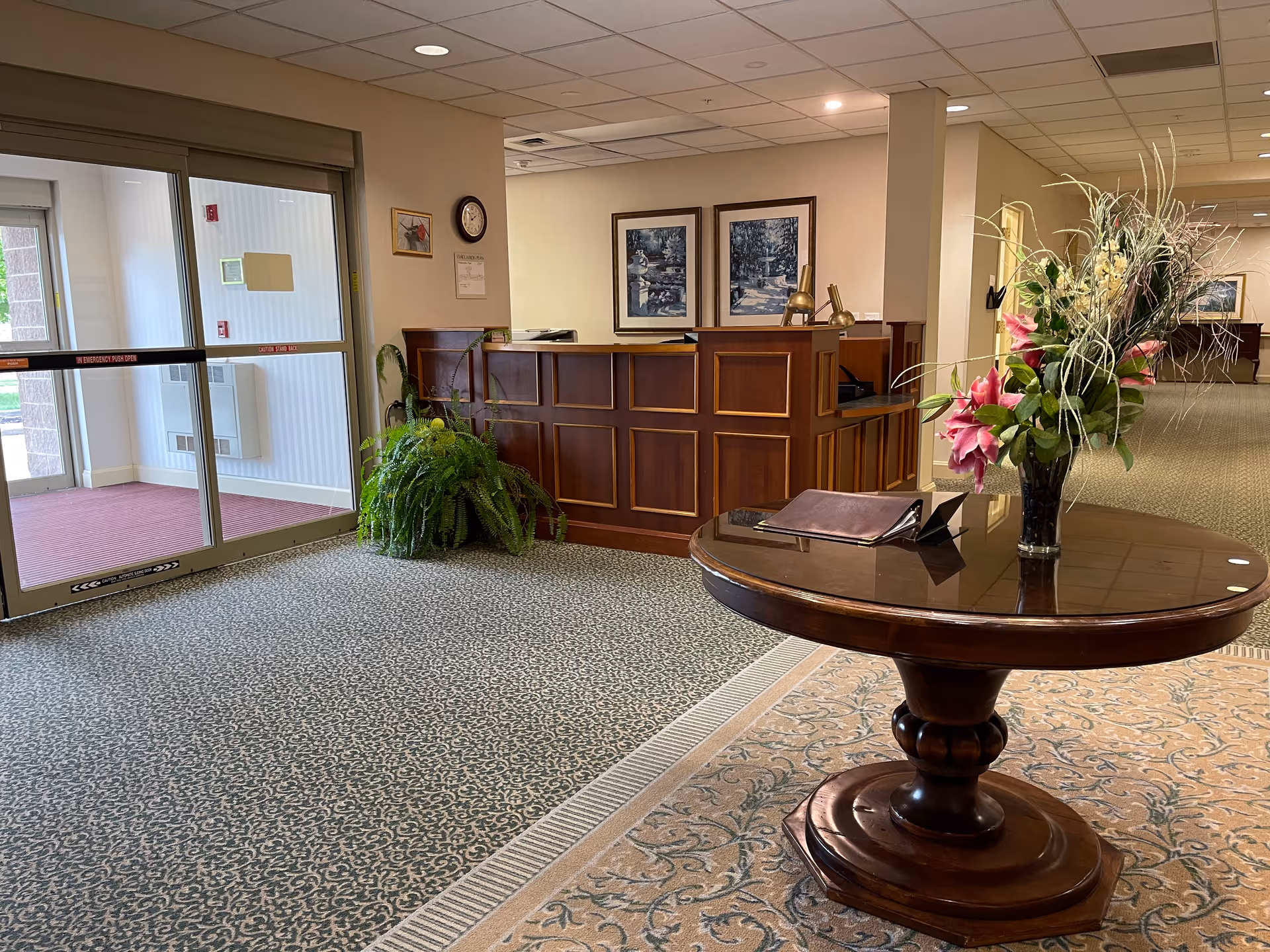 Lobby reception area with a round table topped with flowers in the foreground, a wooden front desk, and glass entry doors.