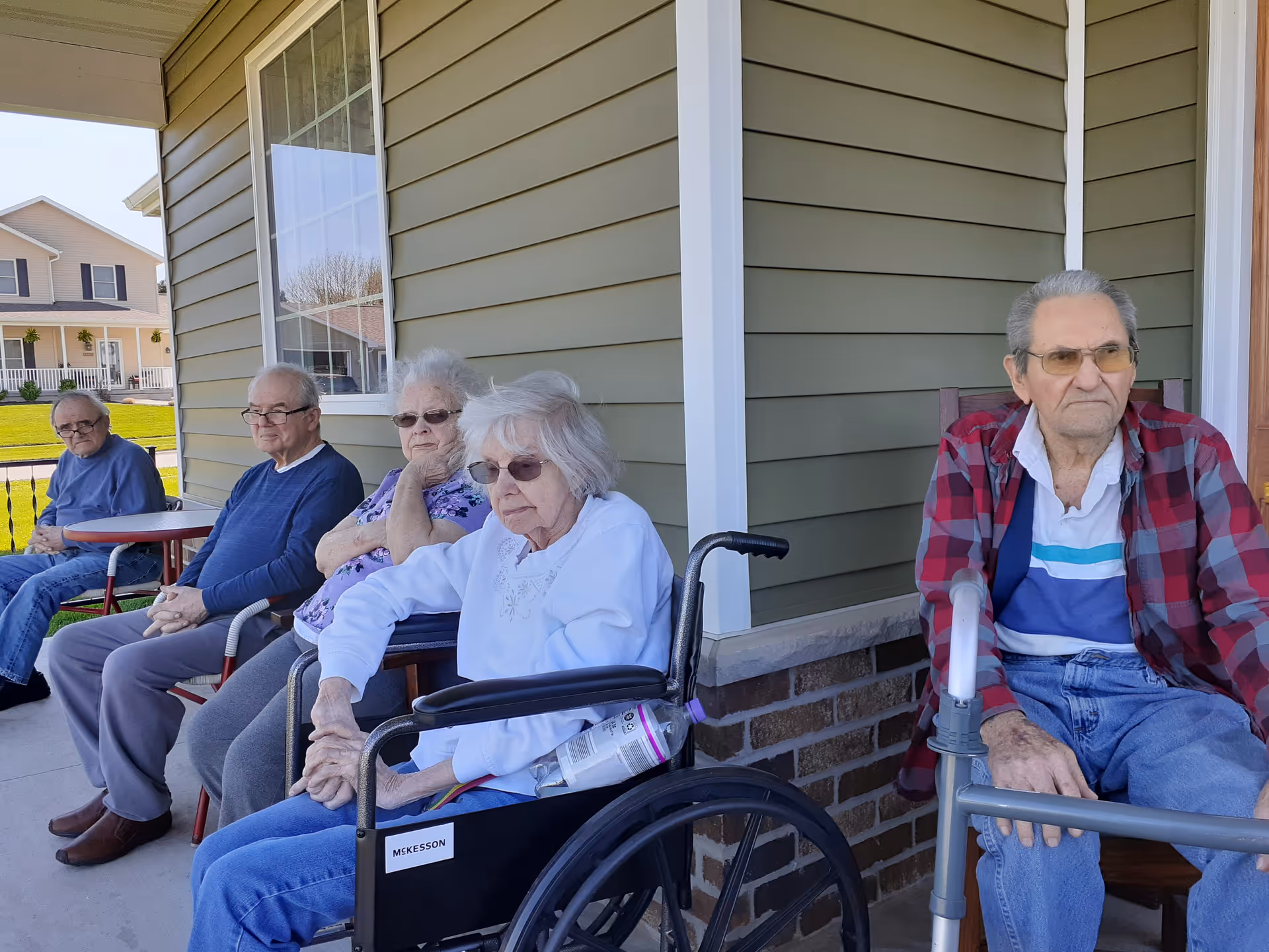 Five elderly individuals sitting outside on a porch area of a building. Two men and three women are seated, with one woman in a wheelchair and one man using a walker. The building has green siding and white trim, and a residential neighborhood is visible in the background.