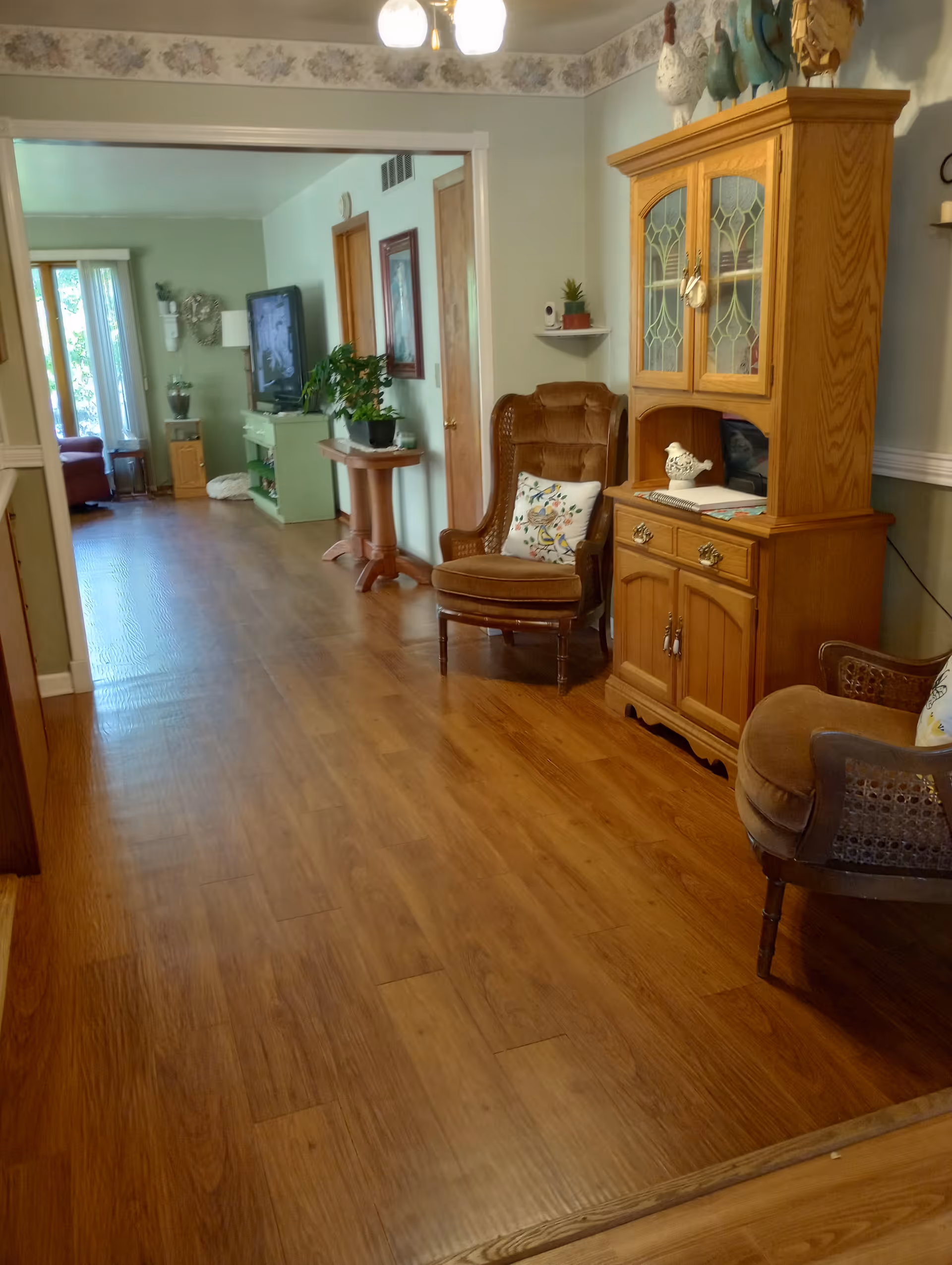 Interior view of a living room hallway with wooden floors, a wooden cabinet, armchairs and a TV visible in the background.