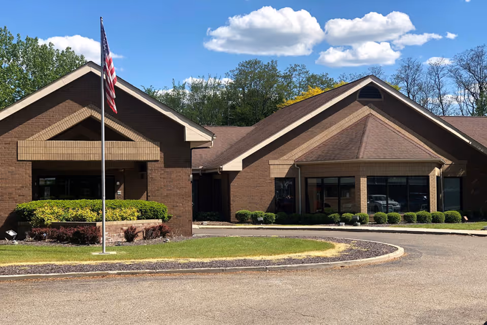 Exterior view of a single-story brick building with a peaked roof, an American flag on a flagpole in front, and neatly trimmed bushes along the building's facade under a blue sky with scattered clouds.