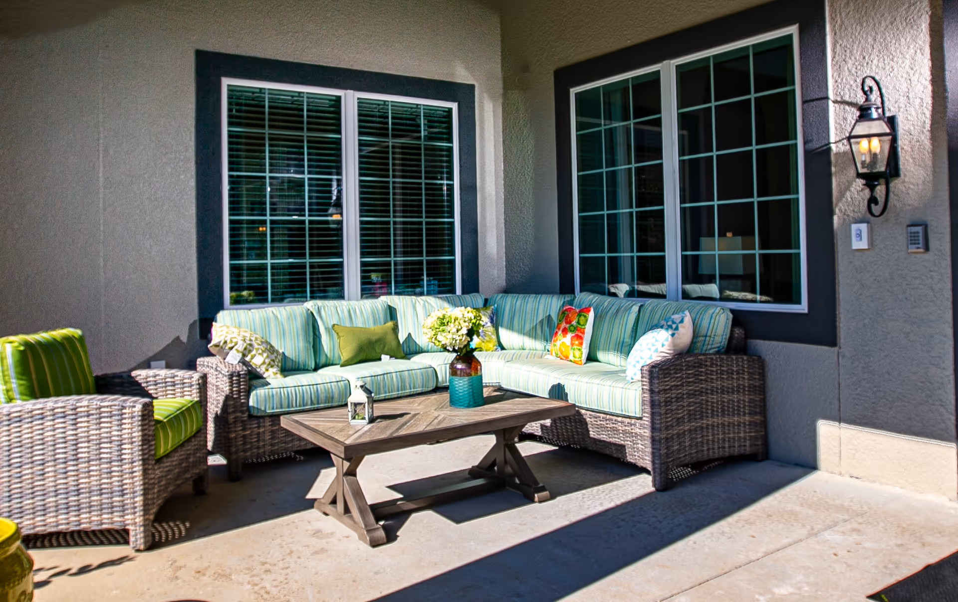 Covered outdoor patio with a wicker sectional sofa with green striped cushions and a wooden coffee table in front of windows.