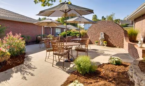 Sunlit outdoor courtyard with patio tables and umbrellas, metal chairs, brick buildings, and landscaped planters.