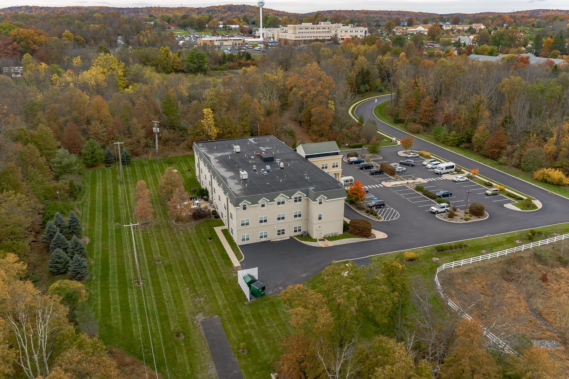 Aerial view of a three-story senior living facility named Hidden Meadows On the Ridge surrounded by green lawns, trees with autumn foliage, a parking lot with several cars, and a winding road leading to the building.