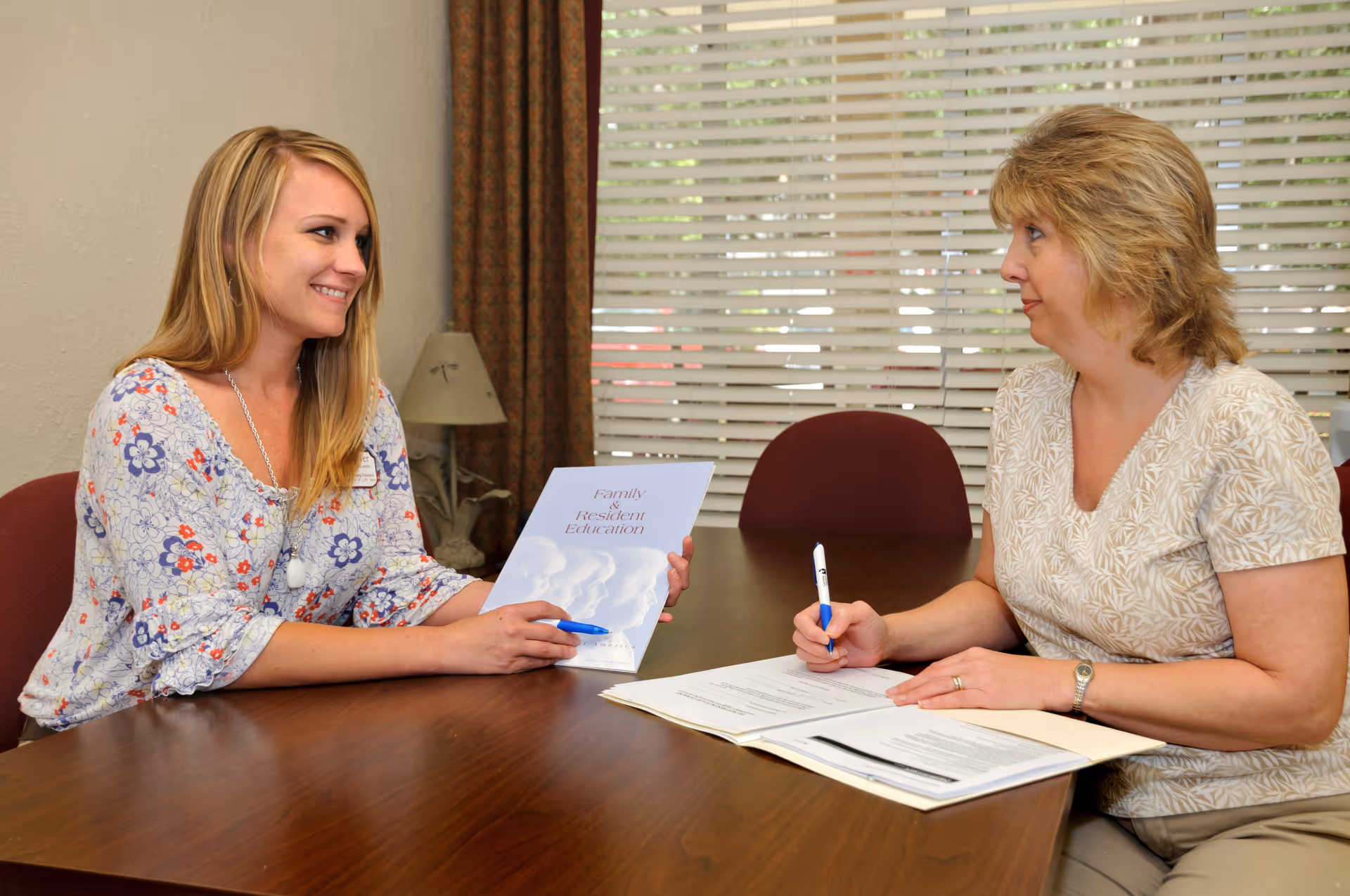 Two women sitting at a wooden table in a room with closed blinds. One woman is holding a booklet titled 'Family & Resident Education' and smiling, while the other woman is writing on a document with a pen.