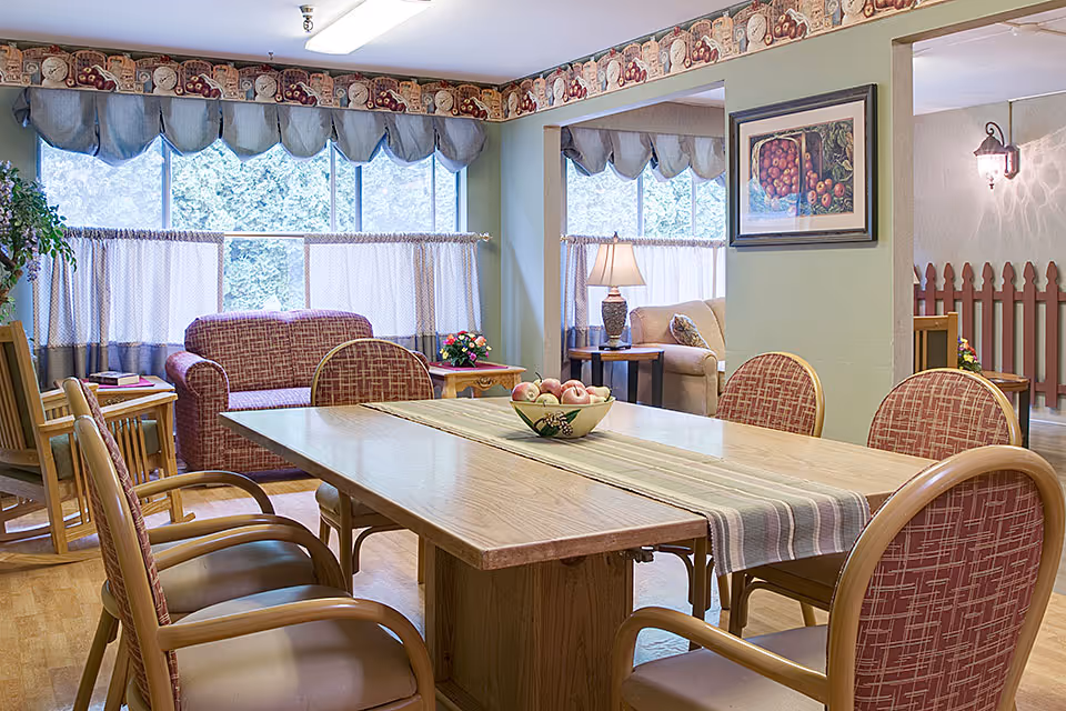 A cozy common area in a senior living facility featuring a wooden table with six cushioned chairs around it. The table has a striped runner and a bowl of apples as a centerpiece. In the background, there are two upholstered couches, a side table with a lamp, large windows with curtains, and decorative wallpaper border near the ceiling.