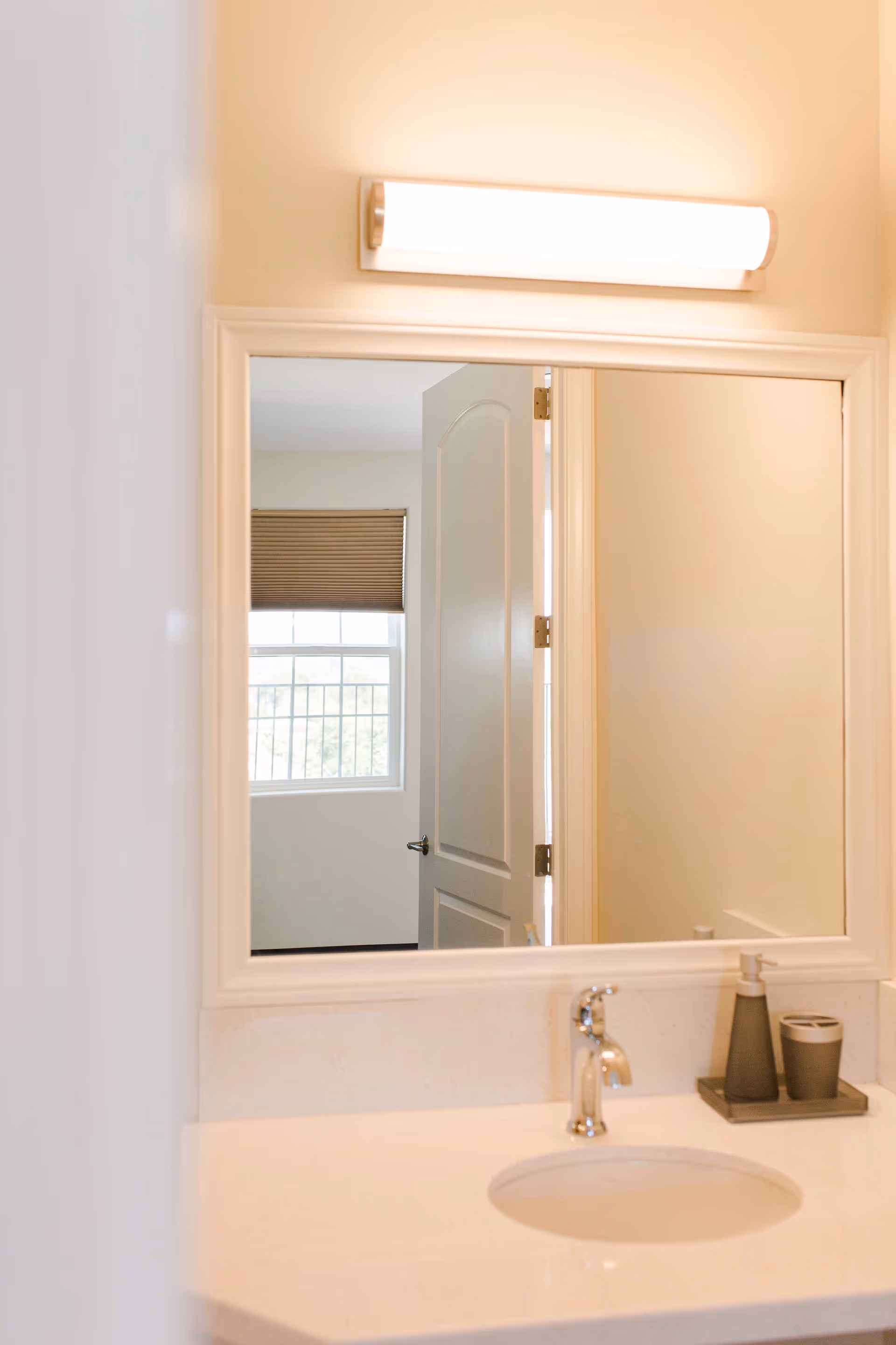 A bathroom sink with a modern faucet, soap dispenser, and cup on a tray. Above the sink is a large rectangular mirror reflecting a partially open door and a window with closed blinds.
