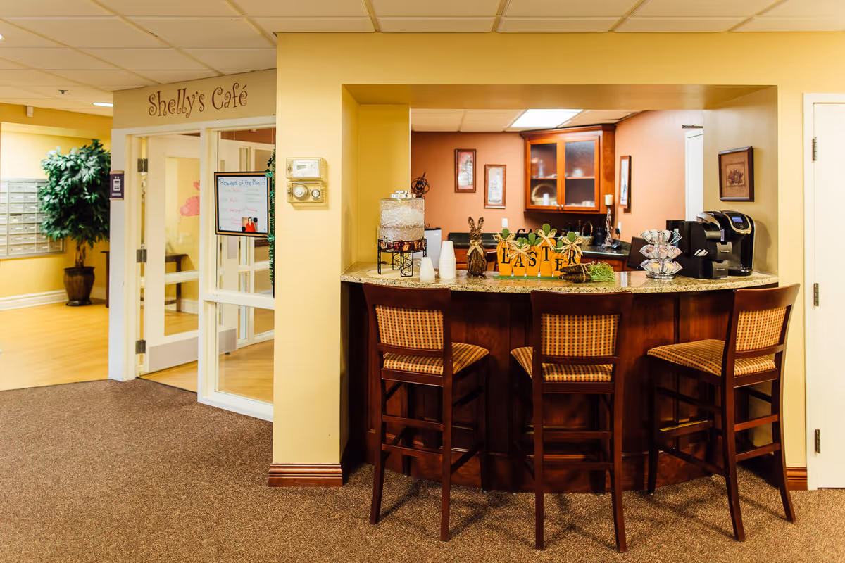 Interior view of a small café area named Shelly's Café with a counter and three wooden bar stools. On the counter, there is a water dispenser, a coffee machine, and Easter-themed decorations. Behind the counter, there are cabinets and framed pictures on the wall. To the left, there is a glass door entrance to the café and a potted plant near a wall with mailboxes.
