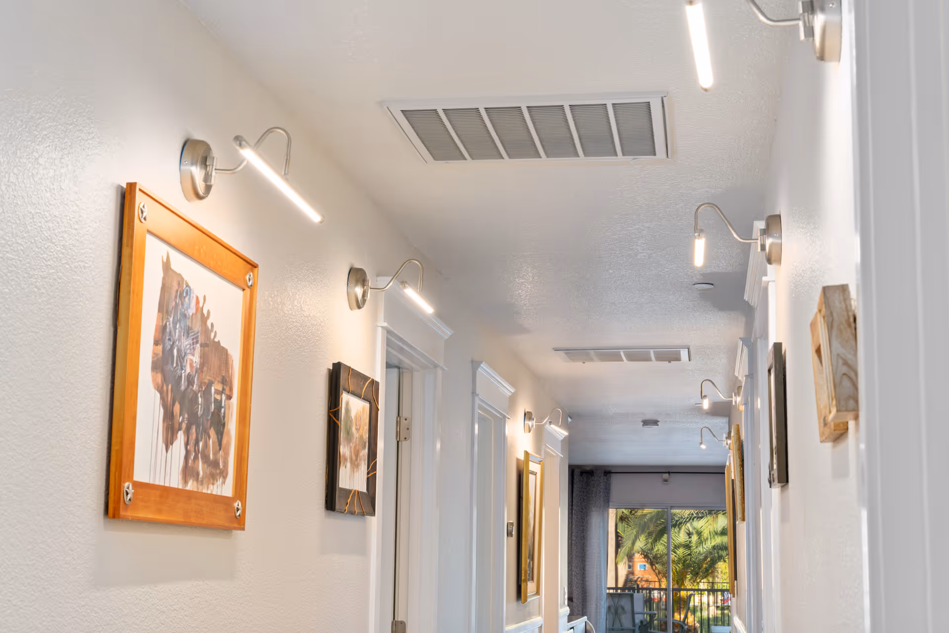 A well-lit hallway with white walls adorned with framed artwork and modern wall-mounted lights. The hallway leads to a glass door with a view of greenery outside.