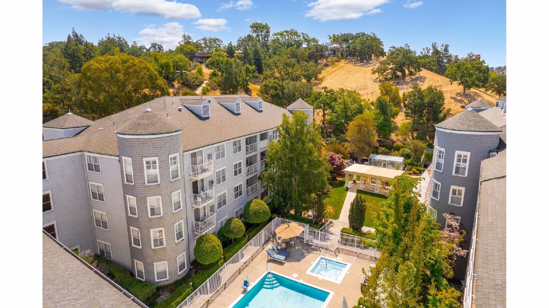 Aerial view of Byron Park senior living facility showing a rectangular swimming pool with adjacent hot tub surrounded by lounge chairs and an umbrella. The pool area is enclosed by a fence and is situated between two multi-story gray buildings with rounded turrets. Behind the pool area is a green lawn with trees, shrubs, and a covered seating area. Hills with trees and dry grass are visible in the background under a partly cloudy sky.