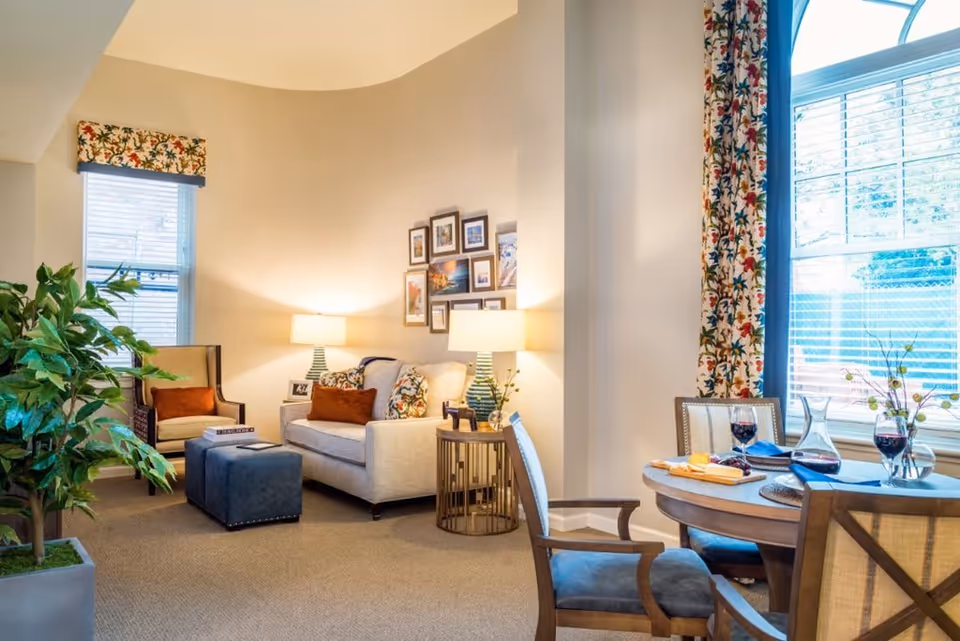 A cozy living room area with a beige sofa adorned with patterned and solid pillows, two table lamps on side tables, a framed photo collage on the wall, a blue ottoman, and an armchair. To the right, there is a round dining table set with wine glasses, a decanter, and napkins, positioned near a large window with floral curtains. A large green potted plant is visible on the left side of the room.