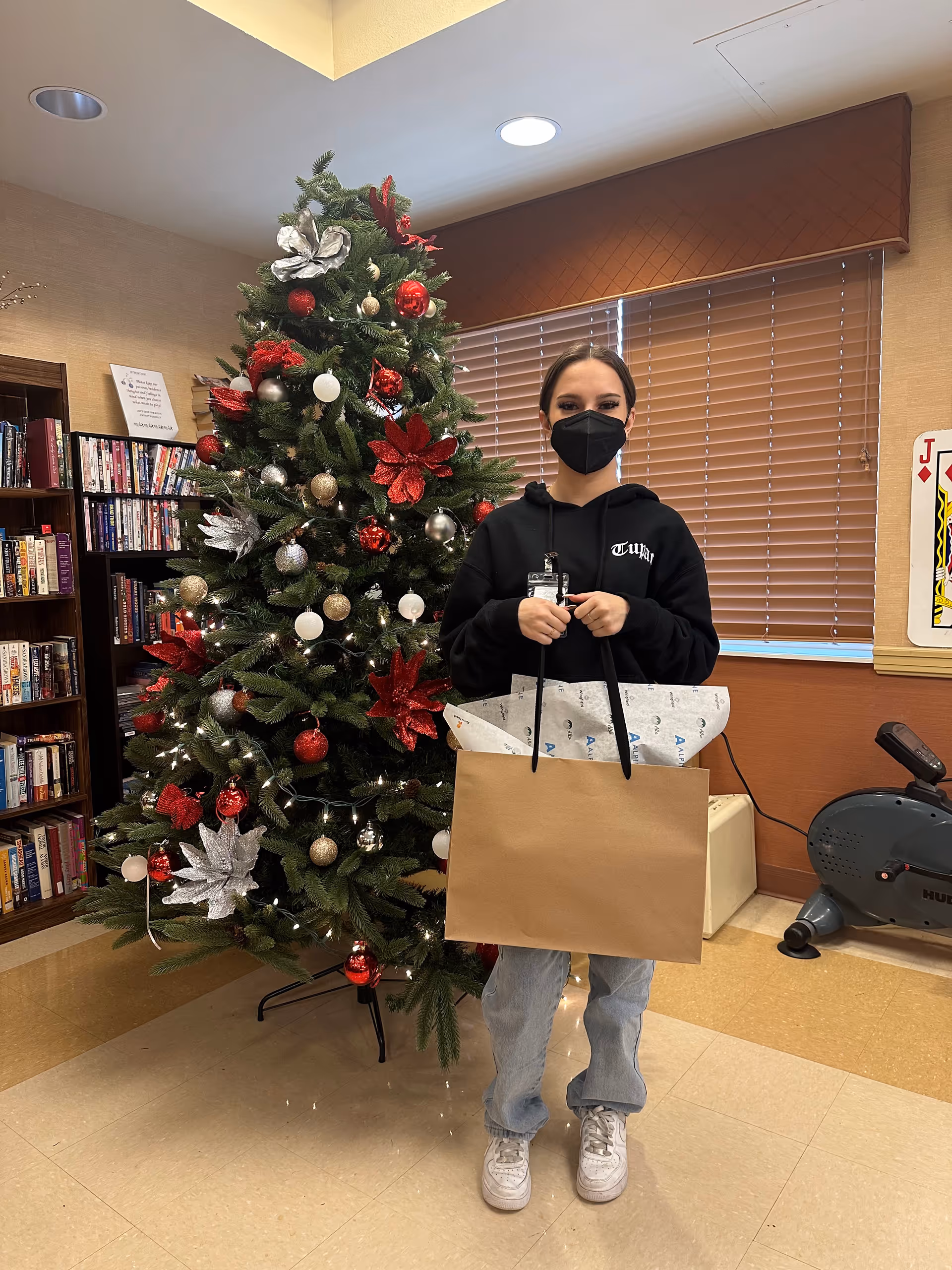 A person wearing a black face mask and black hoodie stands indoors holding a large brown gift bag with tissue paper. Behind them is a decorated Christmas tree with red, silver, and gold ornaments and lights. To the left, there is a bookshelf filled with books, and to the right, a window with closed blinds and an exercise bike are visible.