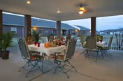 Covered outdoor patio with round dining tables, chairs, potted plants, and ceiling fans at dusk.