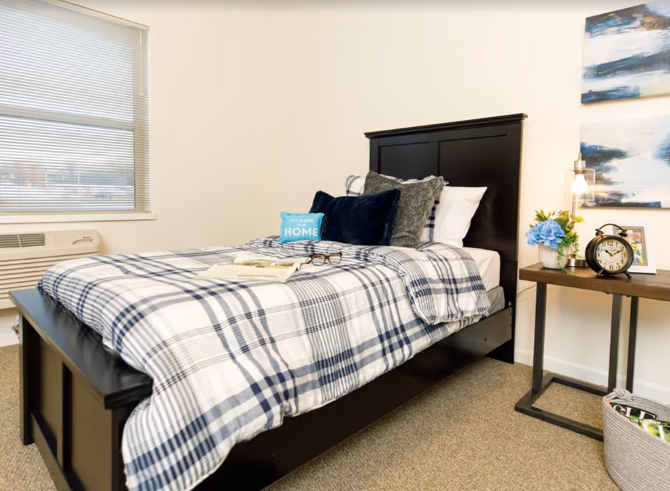 A neatly made single bed with a black wooden frame and plaid bedding in shades of white, gray, and navy blue. The bed has several pillows, including a small blue one with the text 'It's so good to be HOME.' Next to the bed is a wooden side table with a vintage-style alarm clock, a small potted plant with blue flowers, a framed photo, and a modern lamp with an exposed bulb. A window with blinds is visible on the left side of the room, and two abstract paintings hang on the wall above the side table.