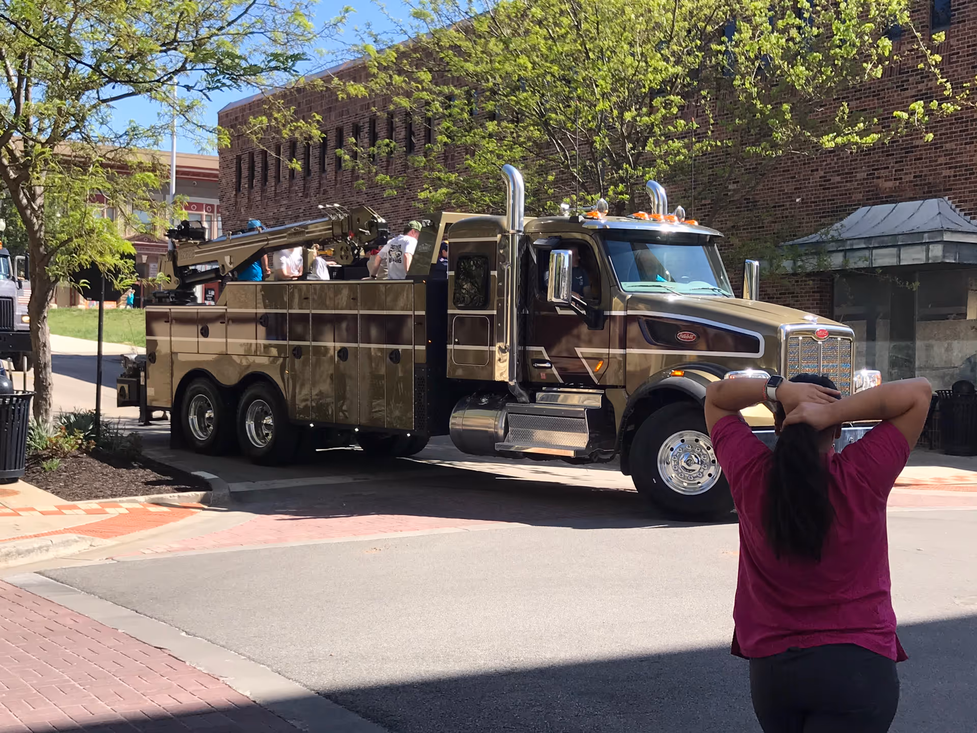 A large tow truck parked on a street with a person in a pink shirt standing in the foreground and a brick building and trees behind.