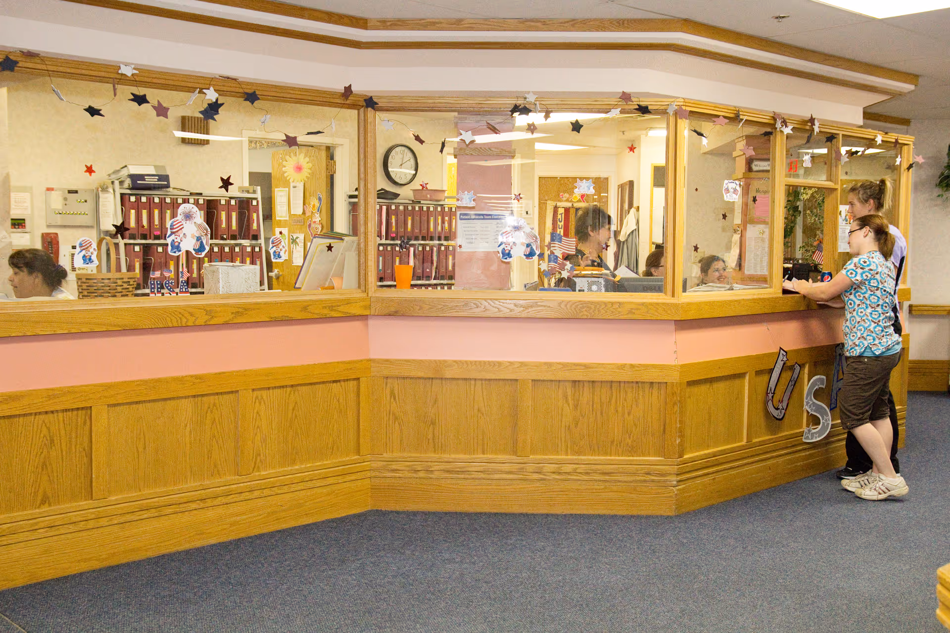 Reception area inside Sidney Health Center Extended Care with a wooden counter and glass partition decorated with star-shaped and patriotic stickers. Two women are standing and interacting with staff behind the counter, where multiple mail slots and office supplies are visible.