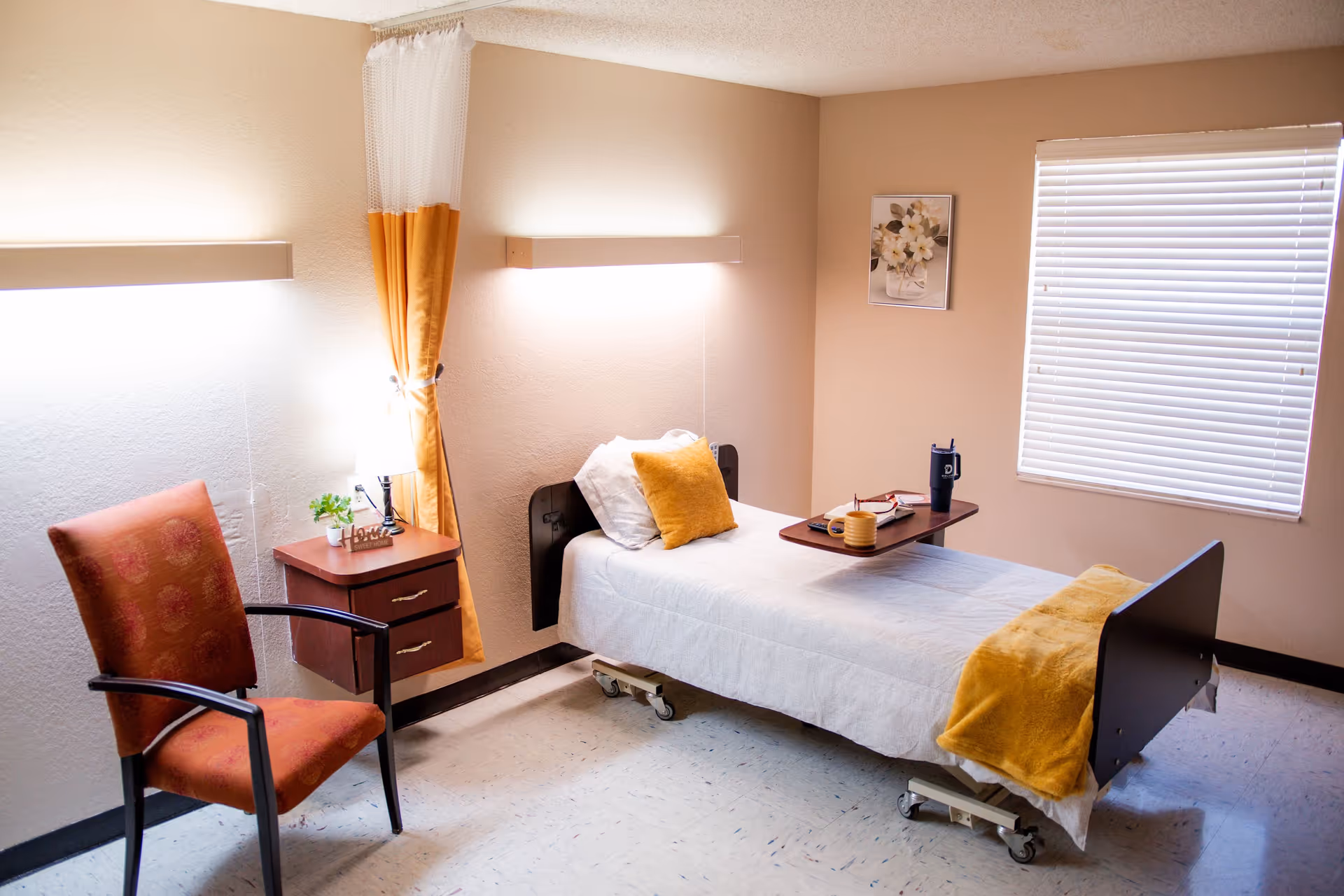 A tidy senior care bedroom with a single bed with yellow accents, a bedside table, chair, and a window with blinds.