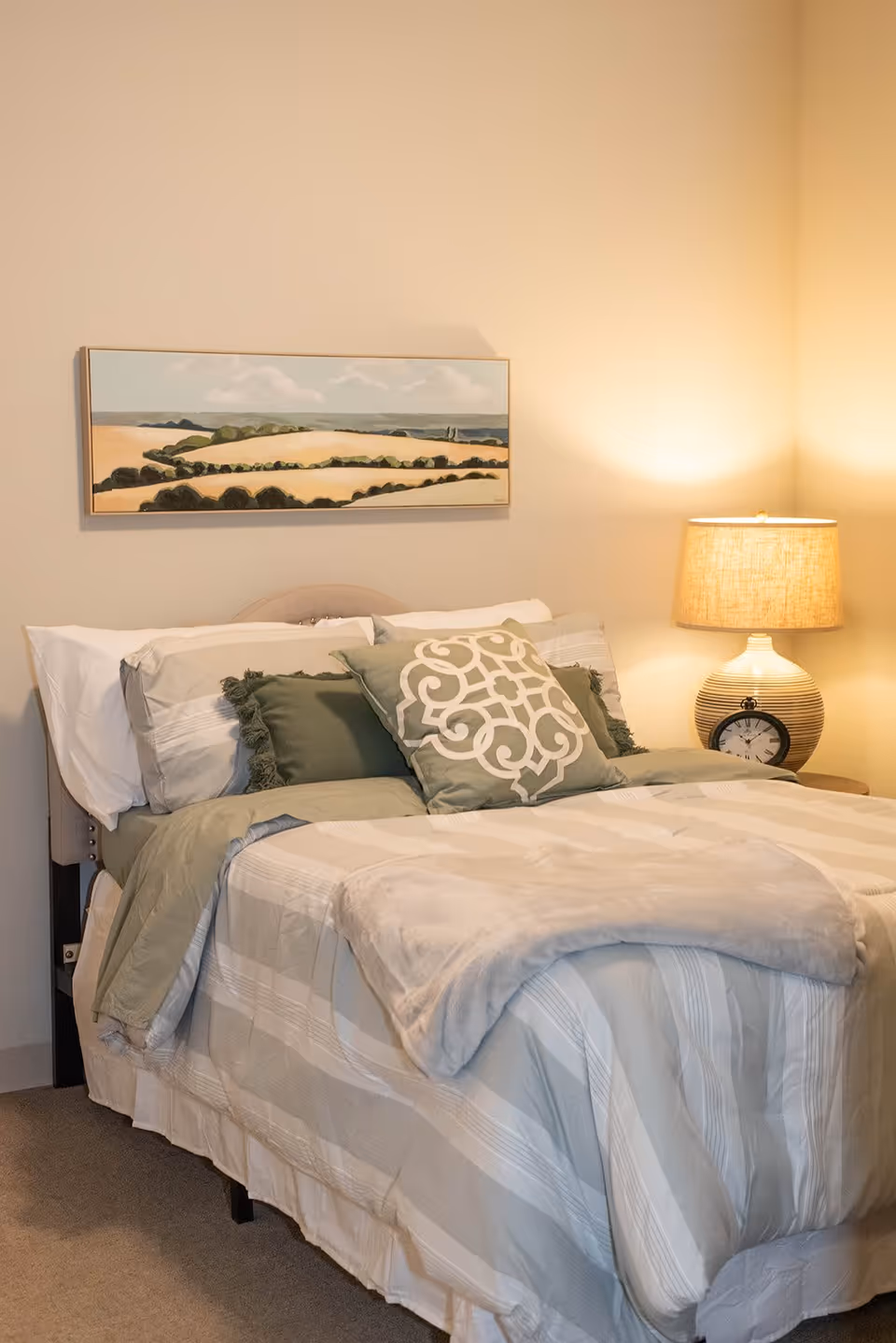 A neatly made bed with beige and green bedding, including decorative pillows, in a softly lit bedroom. A table lamp and a round clock sit on a nightstand to the right of the bed. Above the bed hangs a landscape painting depicting fields and a cloudy sky.