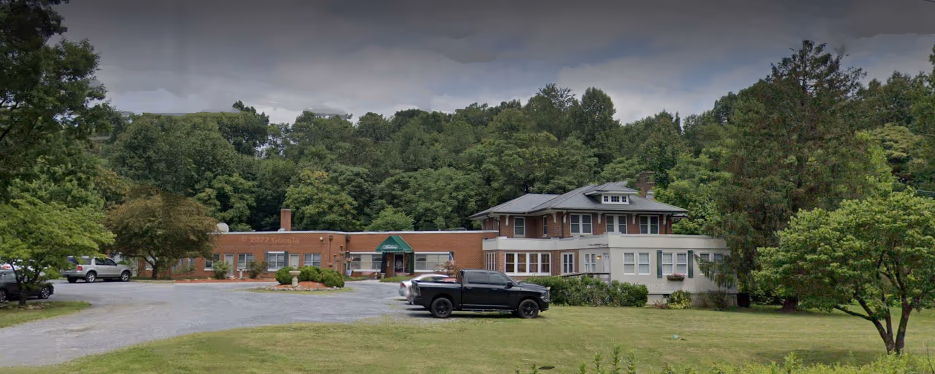 Exterior view of Woodhaven Nursing Home, a single-story brick building with an attached two-story section, surrounded by trees and greenery. Several vehicles are parked in the gravel parking area in front of the building under a cloudy sky.