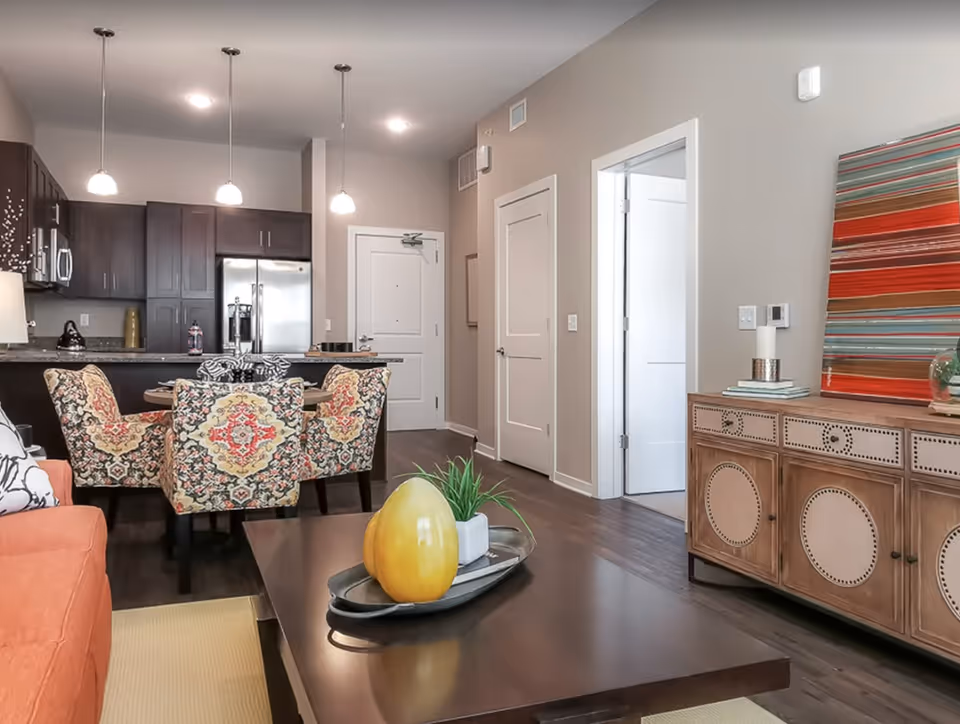 Interior view of a senior living facility's open living and dining area featuring a dark wood coffee table with decorative items, an orange sofa, a dining table with four patterned chairs, and a kitchen with dark cabinets and stainless steel appliances in the background. The walls are painted light beige, and there is a wooden sideboard with a colorful striped artwork above it.