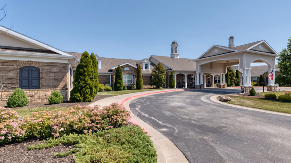 Exterior view of Addington Place of Shoal Creek showing a curved driveway leading to a covered entrance with columns. The building is single-story with brick and stone facade, surrounded by landscaped bushes, trees, and flowering plants under a clear blue sky.