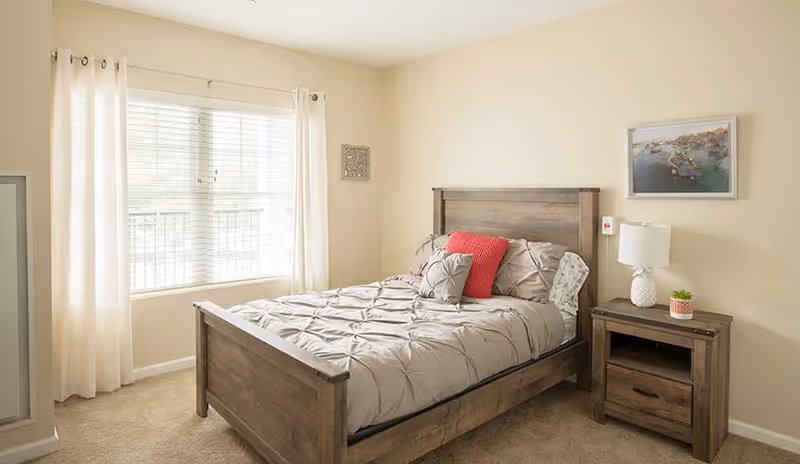 A cozy bedroom with a wooden bed frame and matching nightstand. The bed is made with a light gray comforter and several pillows, including a red accent pillow. A lamp and a small plant sit on the nightstand. There is a large window with white curtains allowing natural light to fill the room, and a framed picture hangs on the beige wall above the nightstand.