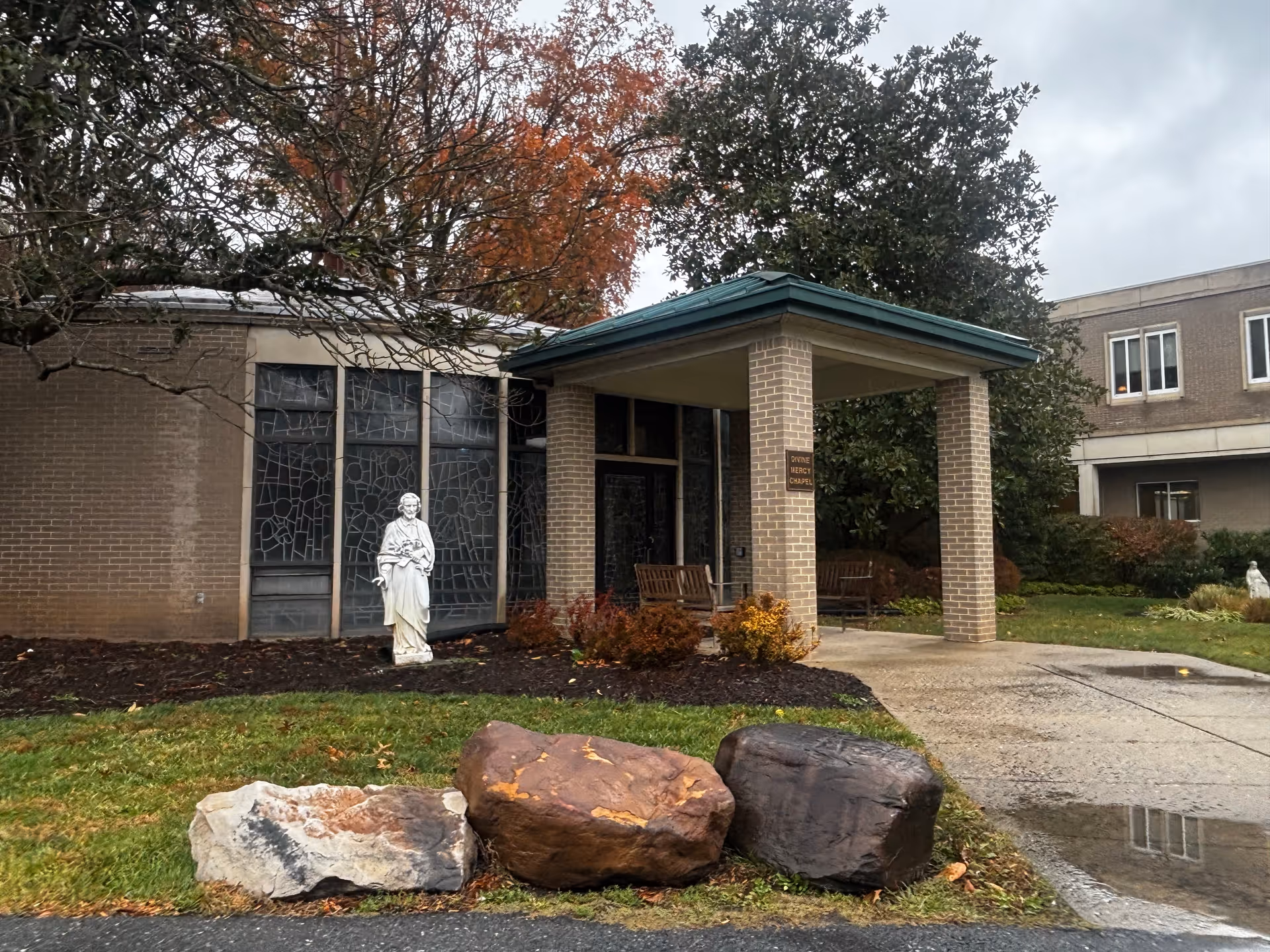 Entrance of a brick senior living facility with a covered portico, a statue by stained glass windows, and large rocks on the lawn.