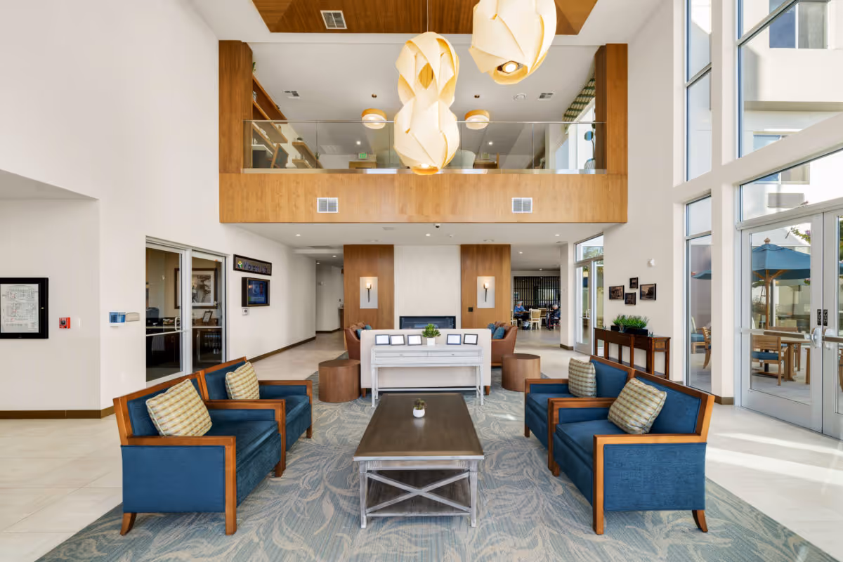 Bright modern senior living lobby with blue armchairs arranged around a coffee table, high ceilings, pendant lights and large windows.