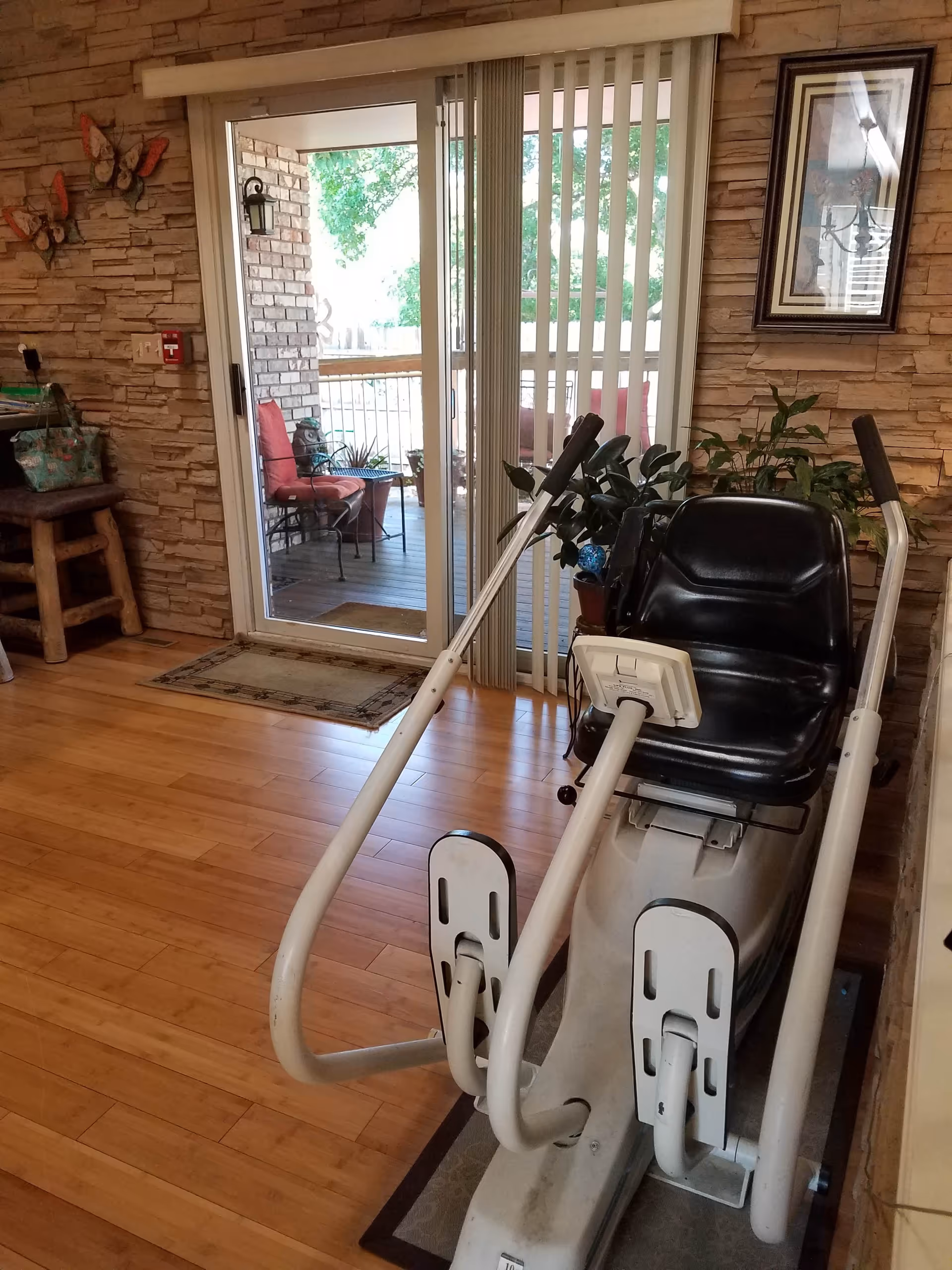 Indoor room with wooden flooring and stone-textured walls featuring a seated exercise machine in the foreground. There is a sliding glass door leading to an outdoor patio with chairs and plants. A framed picture hangs on the wall, and a wooden stool with a bag on it is visible to the left.