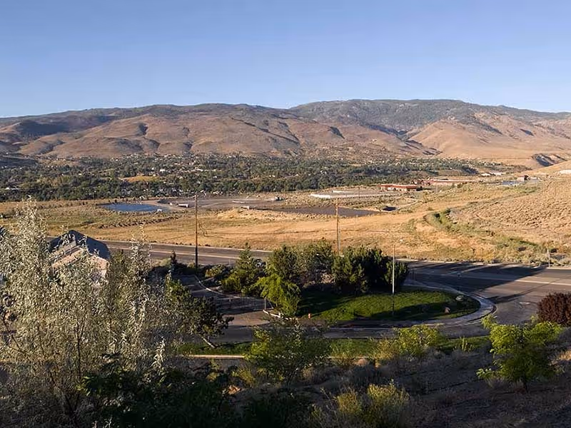 View of a dry, hilly landscape with sparse vegetation and a few trees near a road intersection under a clear blue sky.