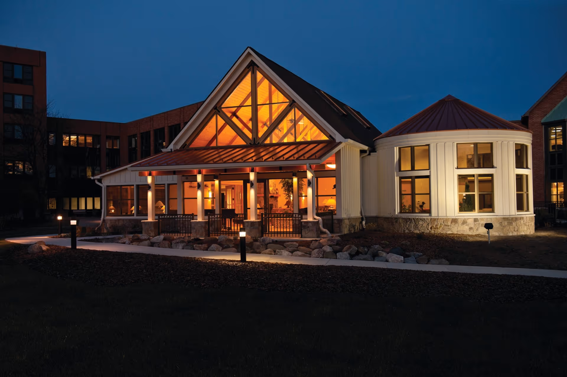 Illuminated entrance and exterior of a senior living building at dusk with a large gabled glass façade and rounded wing.