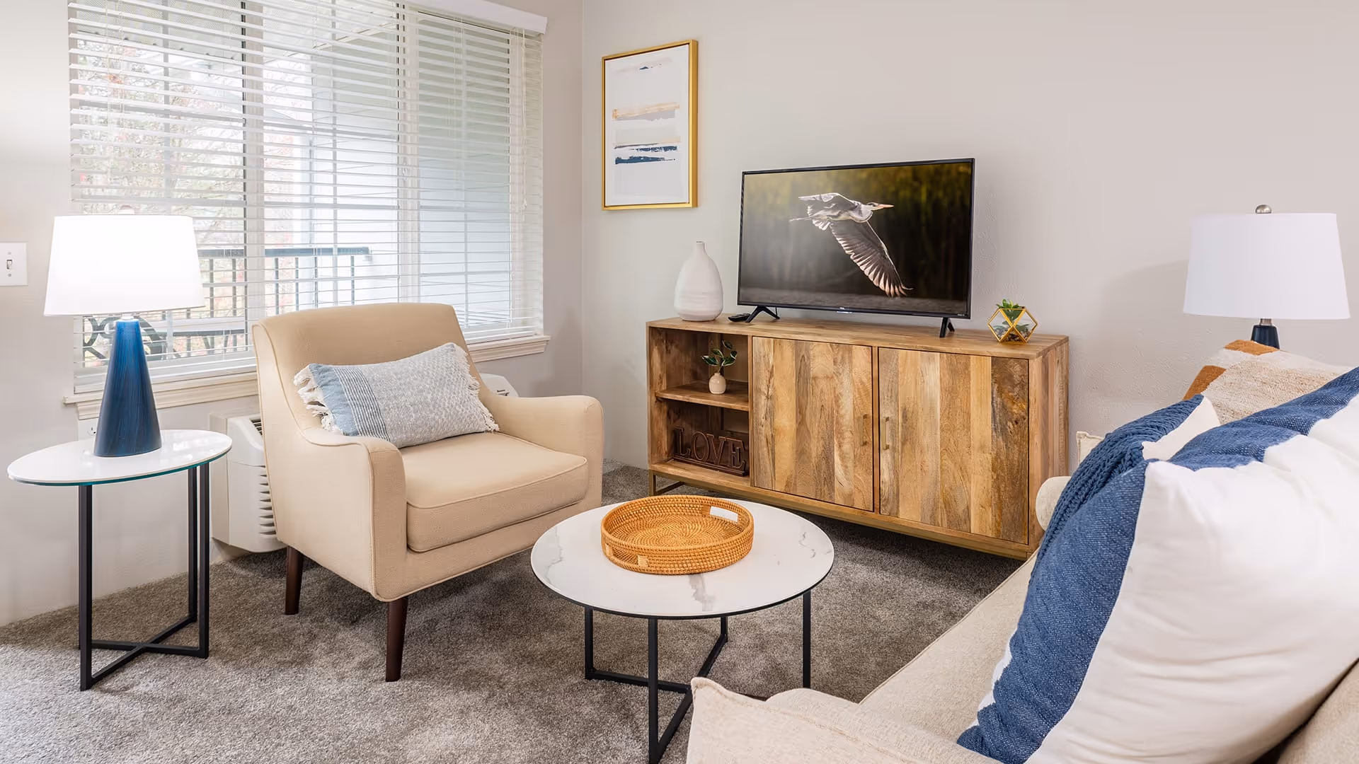 A cozy living room with a beige armchair and a sofa with blue and white pillows. There is a round white marble coffee table with a wicker tray on it, and a wooden TV stand with a flat-screen TV displaying a bird in flight. Two table lamps with white shades are placed on either side of the room, and a large window with white blinds lets in natural light.