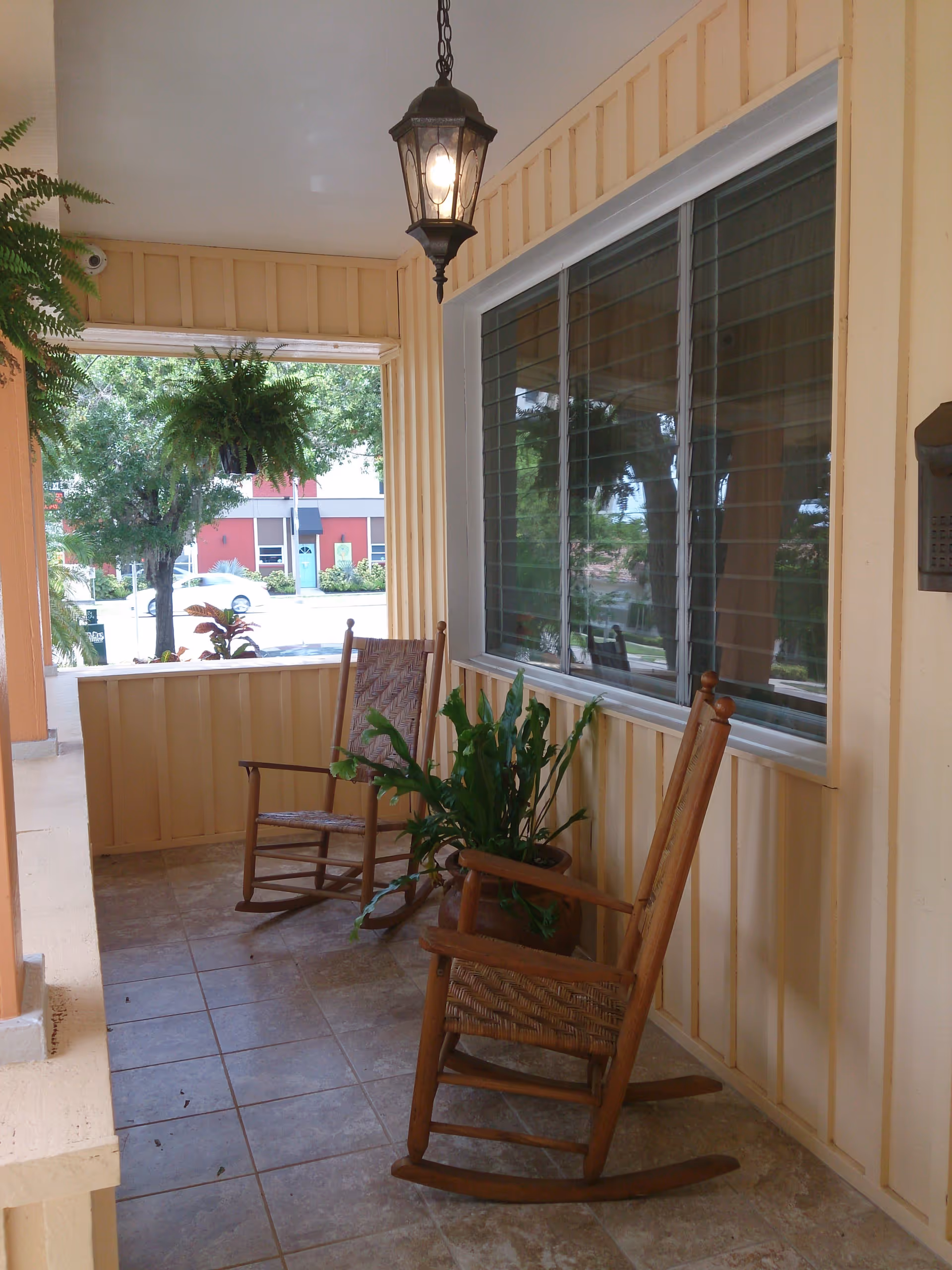 A covered porch area with two wooden rocking chairs and a potted plant between them. There is a hanging lantern light above and a large window with horizontal blinds on the right wall. Outside, trees and a red building are visible.