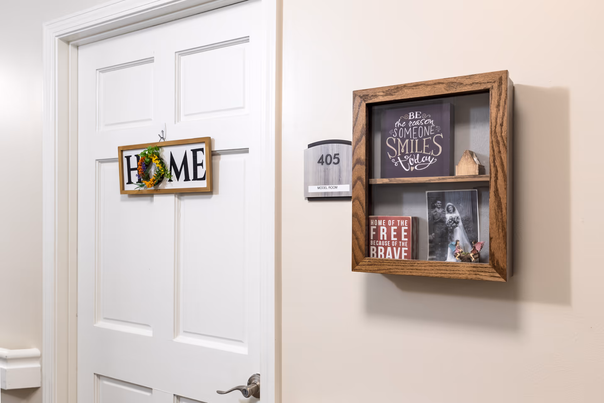 White door with a decorative sign that reads 'HOME' hanging on it, next to a wall-mounted wooden display case containing framed inspirational quotes and a black-and-white photo. A room number plaque labeled '405 MODEL ROOM' is mounted on the wall beside the display case.