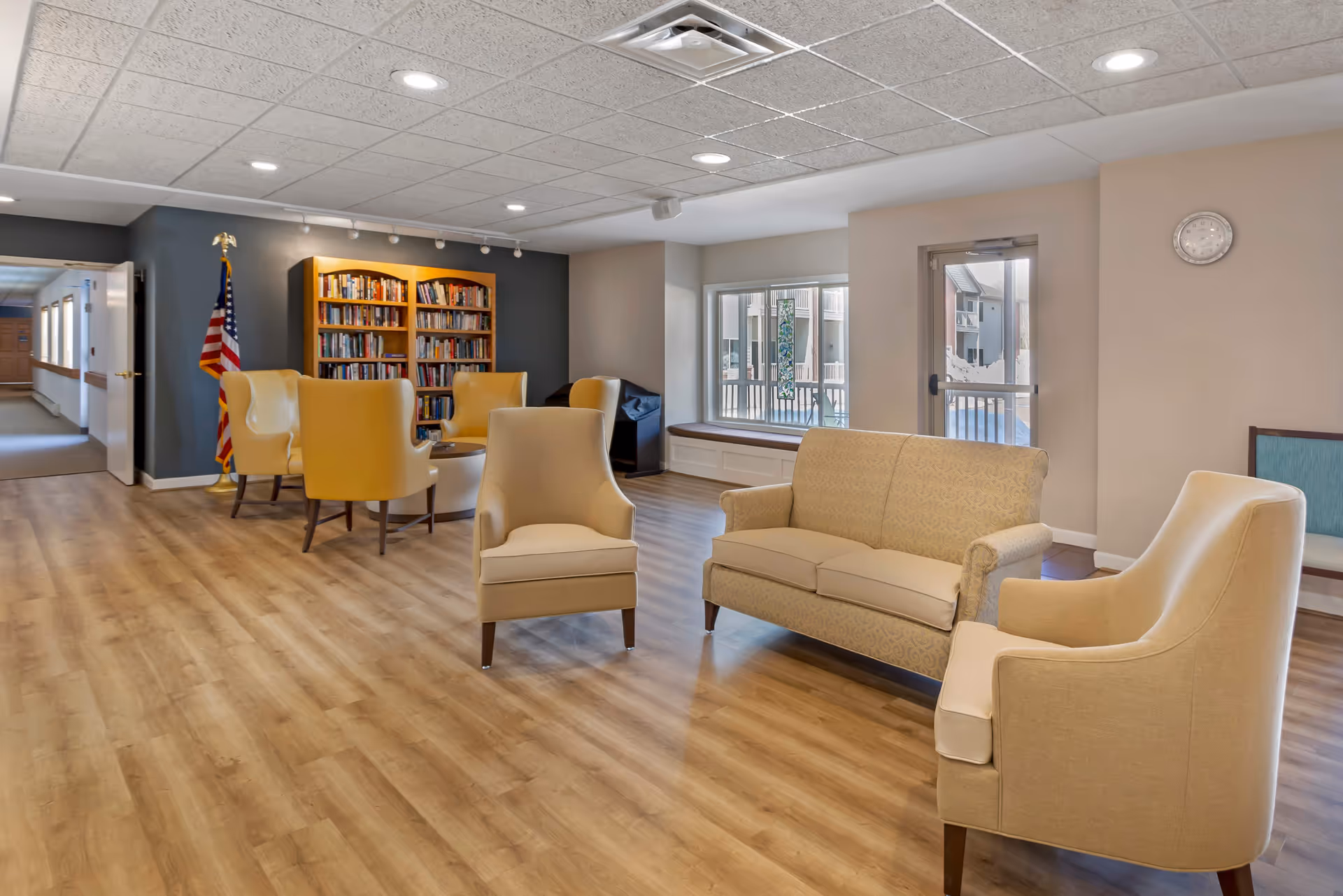 A senior living facility common area with beige and yellow armchairs and a beige patterned sofa arranged on a wood floor. There is a bookshelf filled with books against a dark accent wall, an American flag, a window with a bench seat, and a glass door leading outside. A clock is mounted on a light-colored wall.