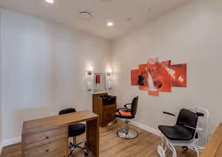 A small room with light-colored walls and wooden flooring, featuring a wooden desk with a black swivel chair, a salon-style chair with a footrest, a small cabinet with a mirror and light fixtures above it, and a multi-panel red and black abstract wall art.