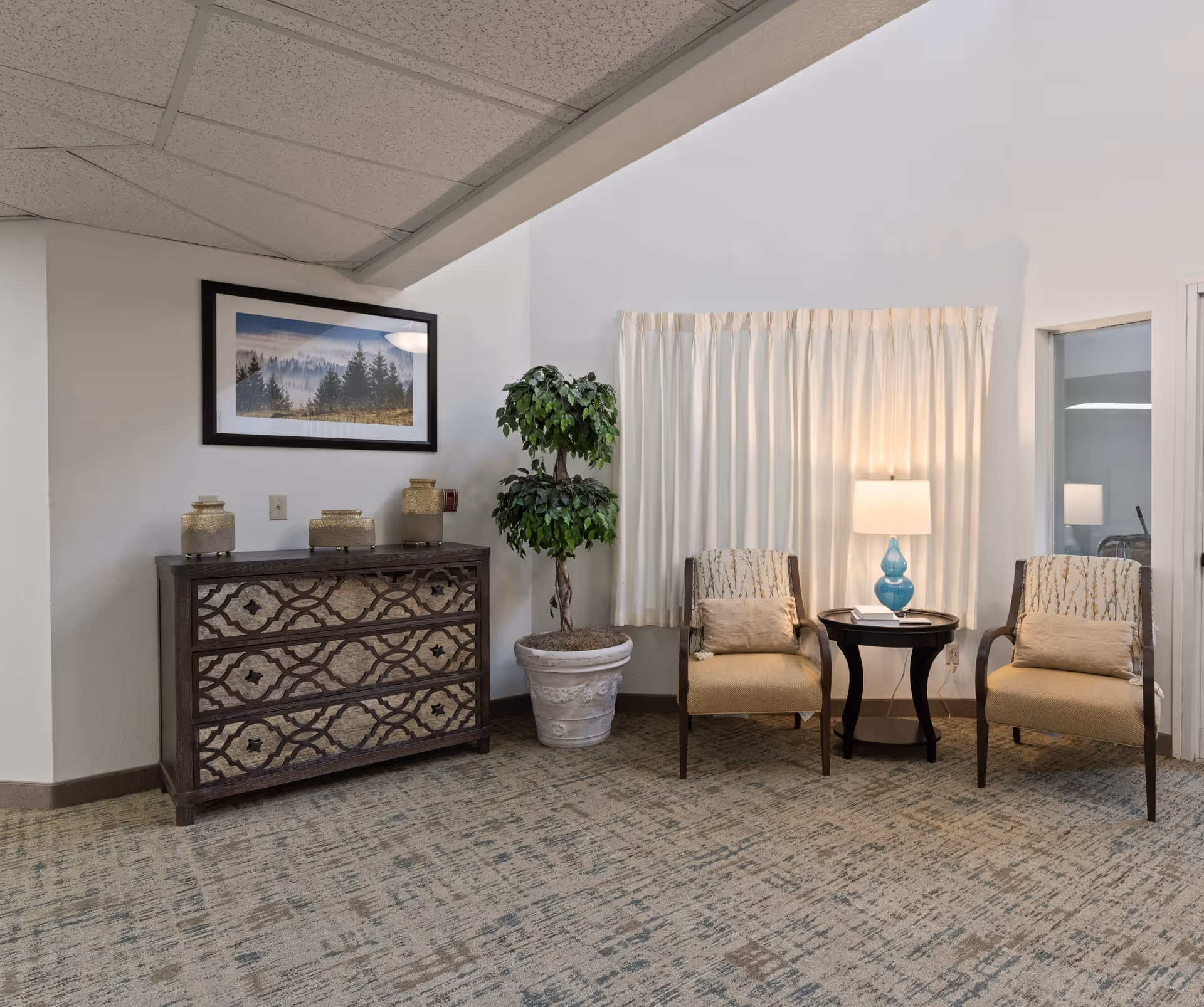 A cozy sitting area in a retirement living facility featuring two beige upholstered chairs with patterned backs and cushions, a small round black side table with a blue glass lamp and a book, a large potted plant, a decorative chest of drawers with three jars on top, and a framed landscape picture on the wall above the chest. The room has light-colored walls, a window with white curtains, and a patterned carpet.
