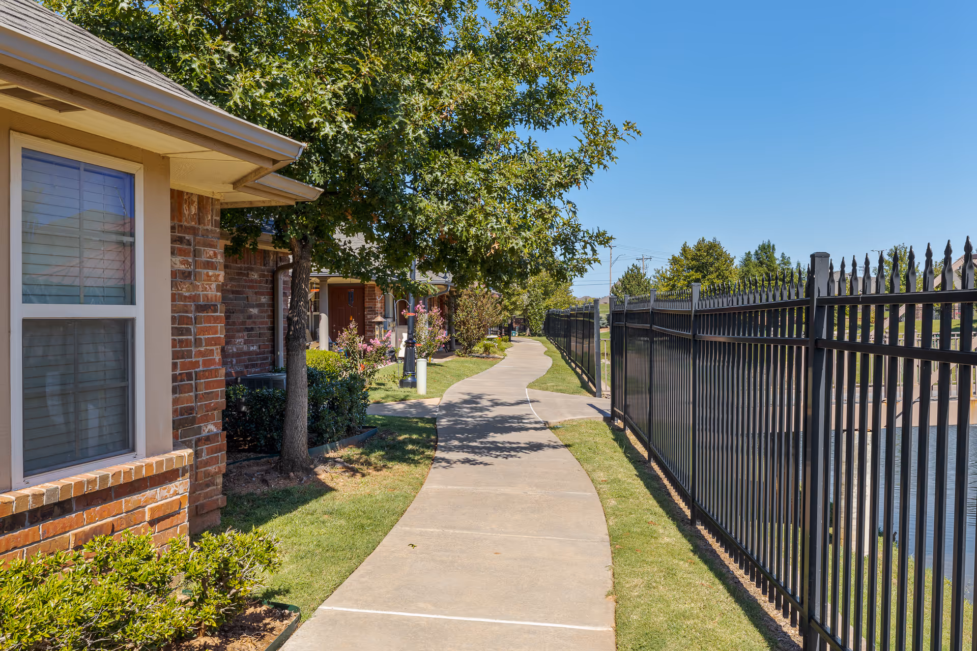 A paved walkway bordered by a black metal fence on the right and a row of brick buildings with windows and trees on the left, under a clear blue sky.