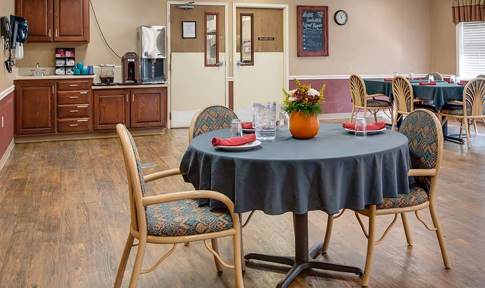 A dining room in a nursing center with round tables covered in dark tablecloths, each set with plates, glasses, and red napkins. One table has a pumpkin vase with flowers as a centerpiece. Wooden chairs with patterned cushions surround the tables. In the background, there is a counter with a coffee machine and supplies, and double doors with windows. A chalkboard menu and a clock are mounted on the wall.
