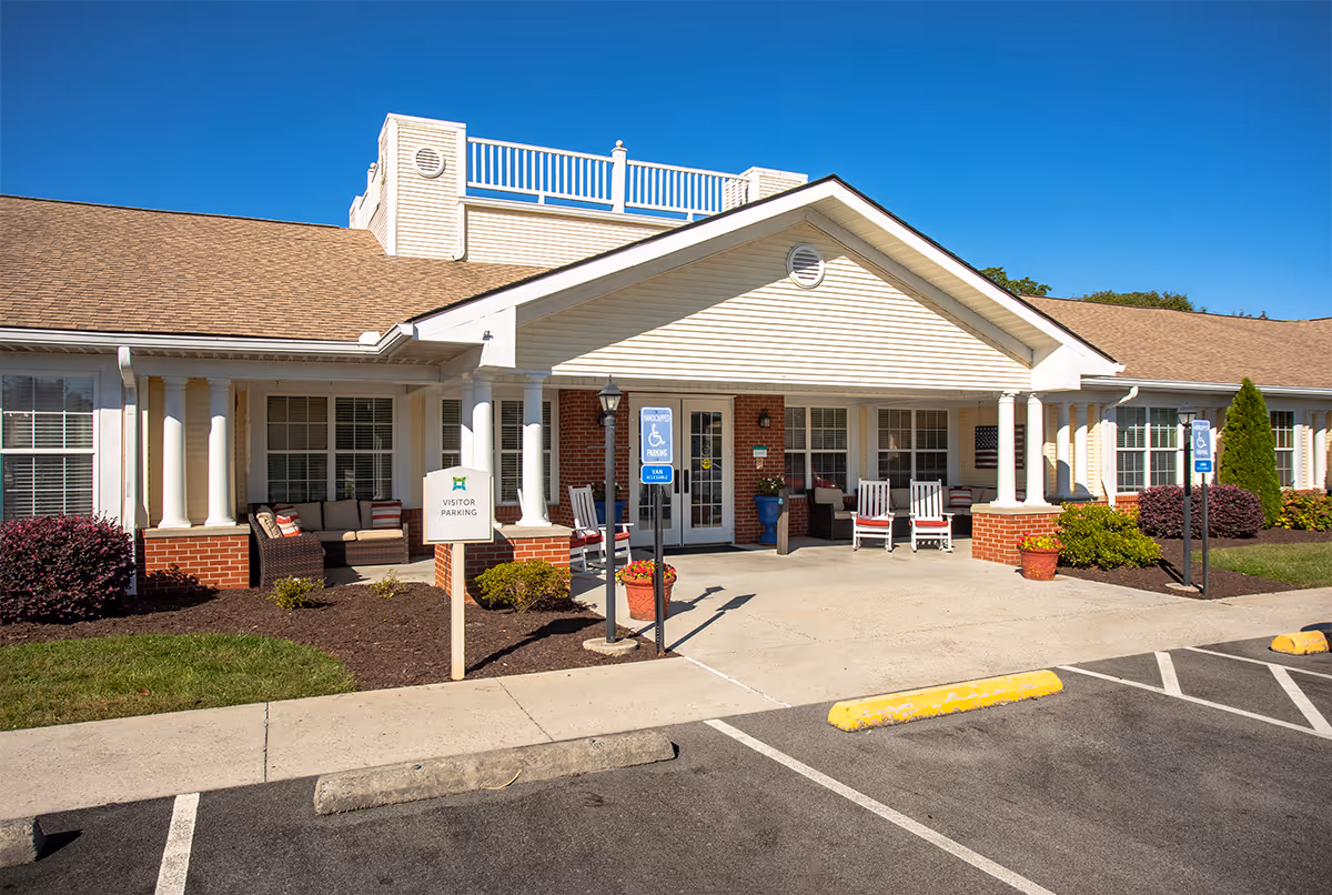 Exterior front view of TerraBella Morristown facility with a covered entrance, white columns, brick accents, and outdoor seating including chairs and a sofa. There are signs indicating visitor parking and handicapped parking spaces in front of the building under a clear blue sky.