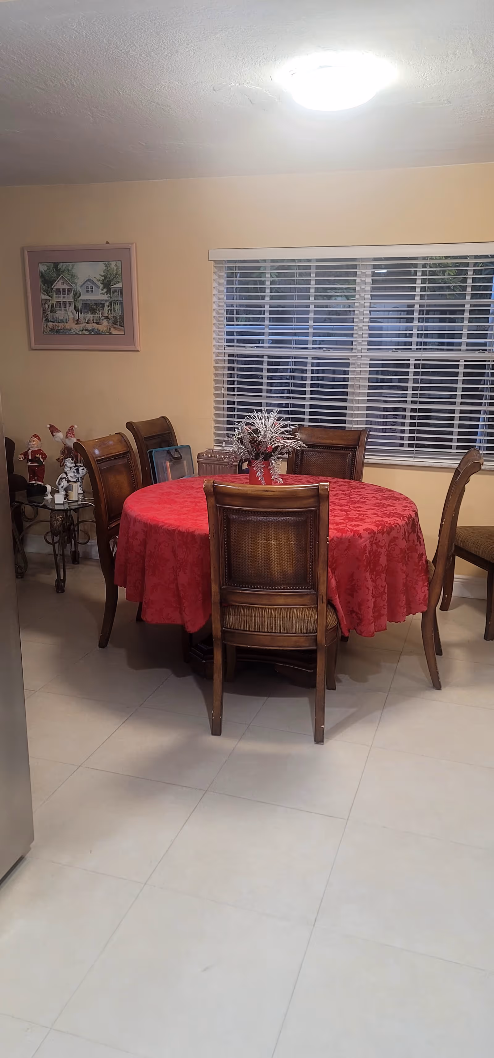 Round dining table covered with a red tablecloth and surrounded by wooden chairs in a room with a window and blinds.