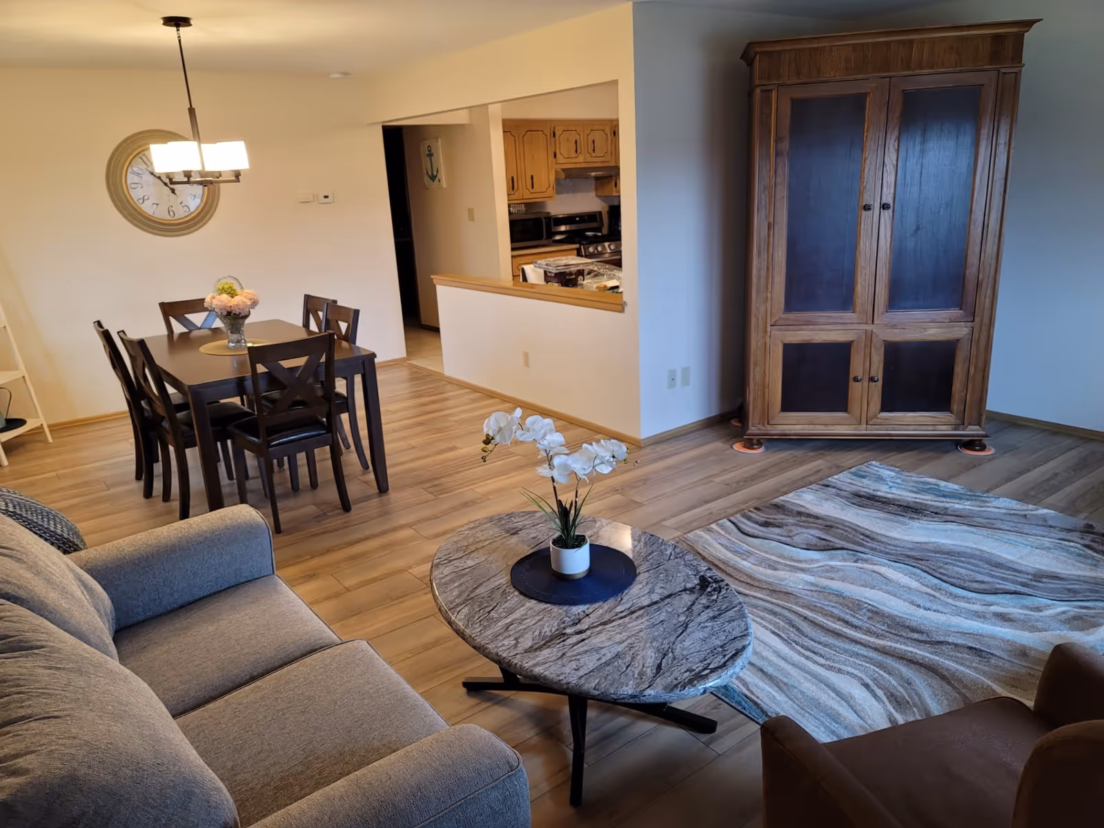 A cozy living and dining area with a gray sofa, a marble coffee table with a white orchid plant, a patterned area rug, a wooden armoire, and a dining table with six chairs under a modern chandelier. The kitchen is visible through a pass-through window.
