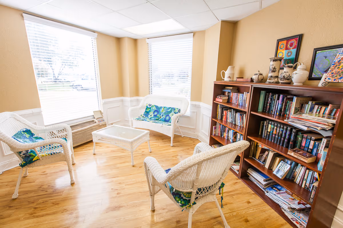 A cozy sitting area with three white wicker chairs and a matching wicker loveseat, all with blue and green floral cushions. A small glass-top wicker table is in the center. The room has light wood flooring, beige walls with white wainscoting, and two large windows with white blinds letting in natural light. To the right, there are two wooden bookshelves filled with books, magazines, and decorative pottery and framed artwork on top.