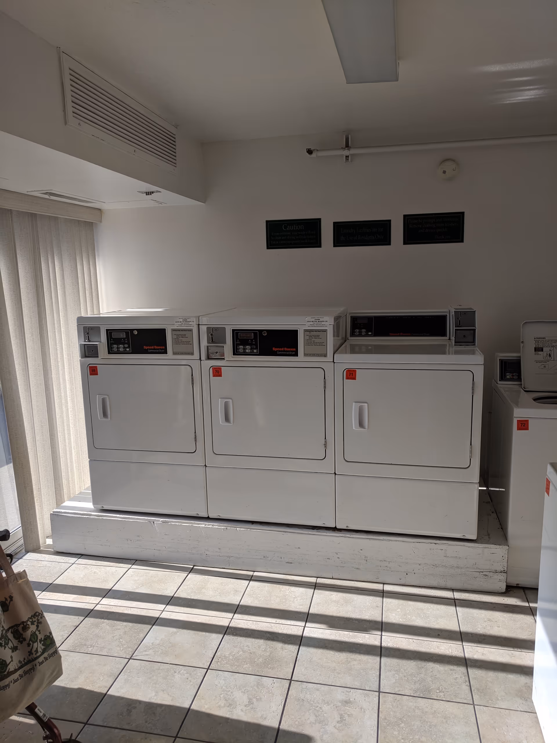 Laundry room with three white commercial dryers labeled 69, 70, and 71, and a washing machine labeled 72. The room has tiled flooring, white walls, and vertical blinds covering a window on the left side. There are three small signs on the wall above the dryers.