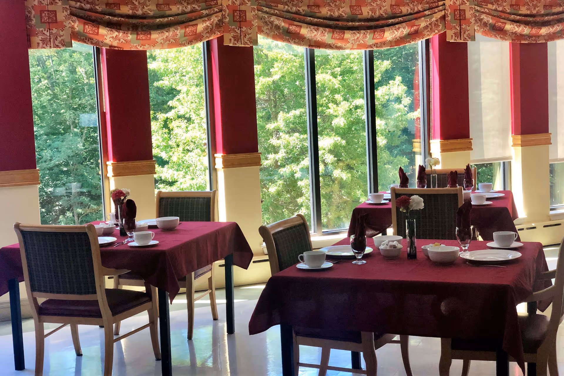 Dining area with tables covered in burgundy tablecloths, set with white plates, bowls, cups, and glasses. Each table has a small flower arrangement. Large windows with patterned valances allow natural light and show green trees outside.