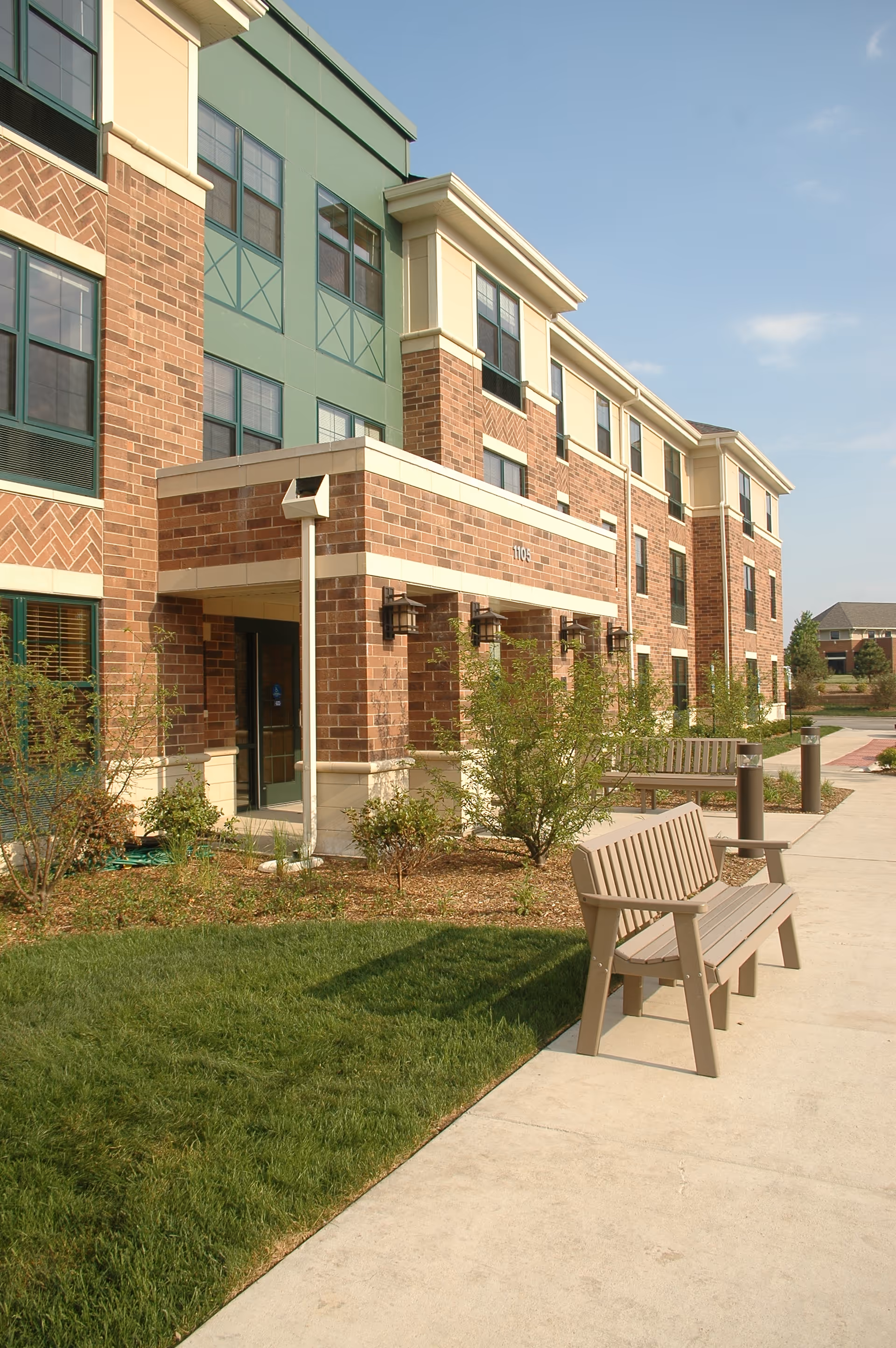 Exterior view of a multi-story brick and green building with a covered entrance, surrounded by landscaped bushes and grass. There are wooden benches along a concrete walkway under a clear sky.