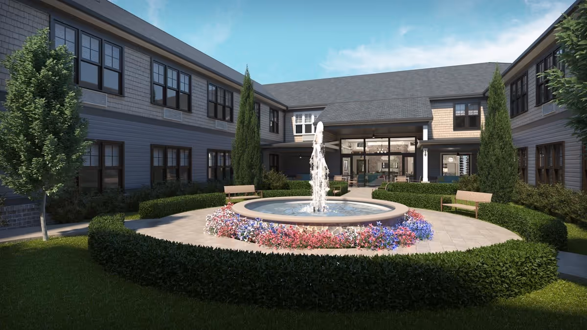 Courtyard with a central fountain surrounded by flower beds, benches, and a two-story senior living building.