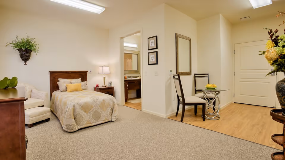 A cozy bedroom in an assisted living facility featuring a single bed with beige patterned bedding, a wooden headboard, a nightstand with a lamp, and a small armchair with an ottoman. There is a small dining area with two chairs and a round glass table with a bowl of fruit. The room has beige carpeting and light-colored walls, with a doorway leading to a bathroom visible in the background.